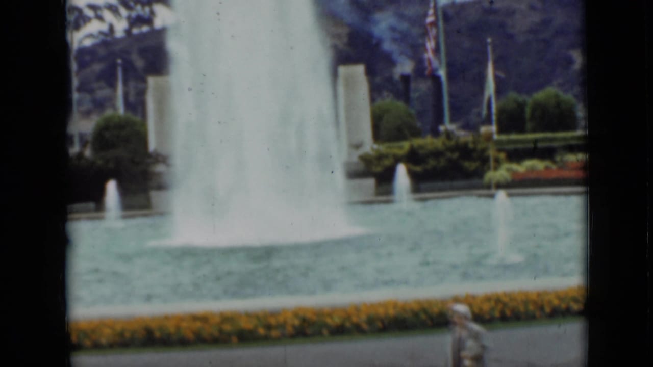1937 San Francisco formal park view with a large fountain plume, smaller jets, yellow flower border, flagpoles, trees, and a person partly visible near the bottom.