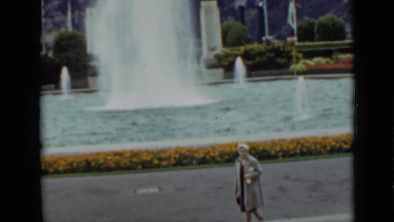 Woman in a light coat and hat pauses beside a fountain pool with flower beds behind her.