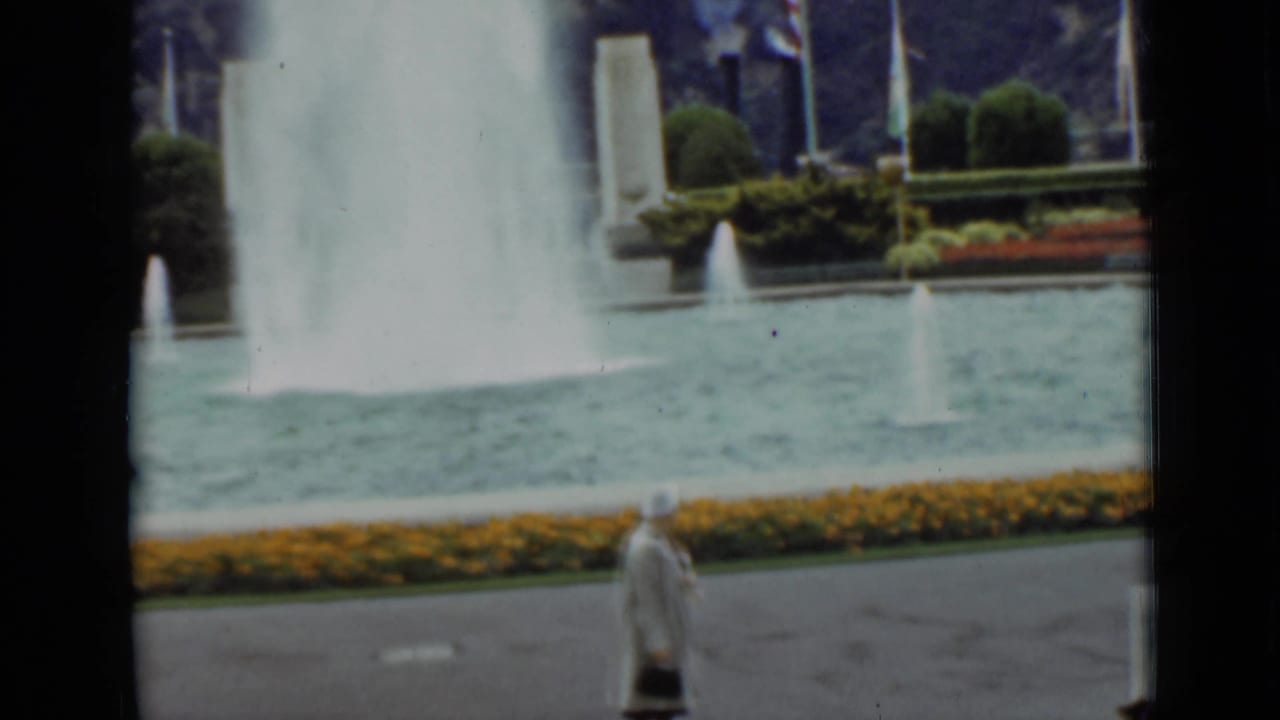 1937 San Francisco frame showing a woman in a light coat moving past a fountain pool bordered by flowers and greenery.