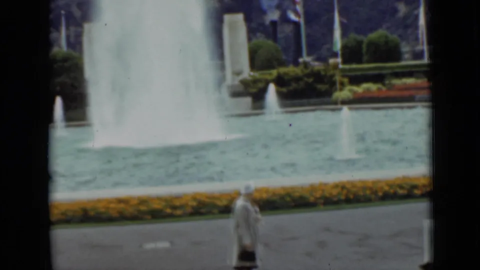1937 San Francisco frame showing a woman in a light coat moving past a fountain pool bordered by flowers and greenery.
