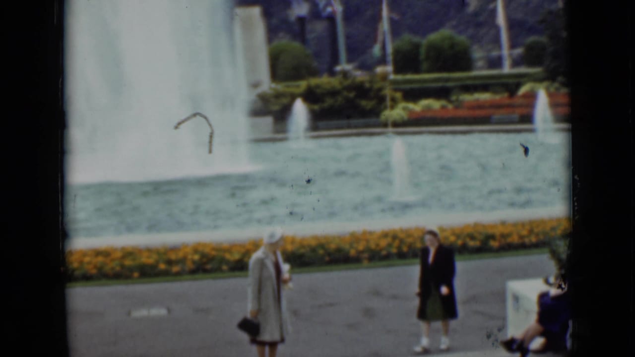 1937 San Francisco home-movie frame with two adult women crossing near a fountain pool and flower border.