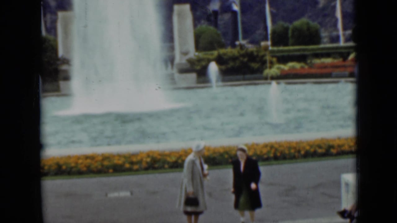 1937 San Francisco frame of two women near a courtyard fountain, with yellow flowers, clipped shrubs, and flags behind the pool.