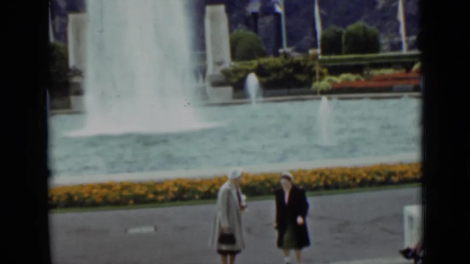 1937 San Francisco frame of two women near a courtyard fountain, with yellow flowers, clipped shrubs, and flags behind the pool.