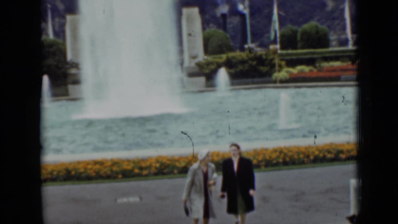 Two women walking near a large fountain and flower bed in a park.