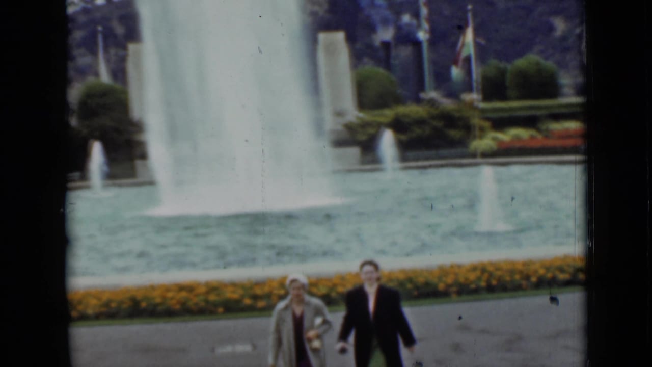 The women continue walking beside the fountain pool in 1937 San Francisco footage, with flower beds, flags, and shrubs in the background.