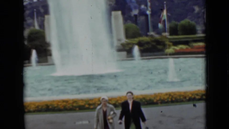 The women continue walking beside the fountain pool in 1937 San Francisco footage, with flower beds, flags, and shrubs in the background.