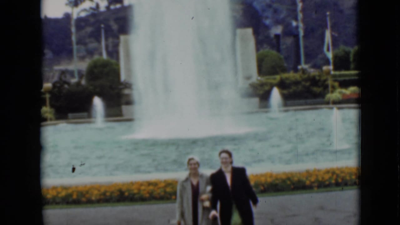 Two women in coats and hats walk in the lower frame while a tall fountain spray rises behind them in the San Francisco park scene.