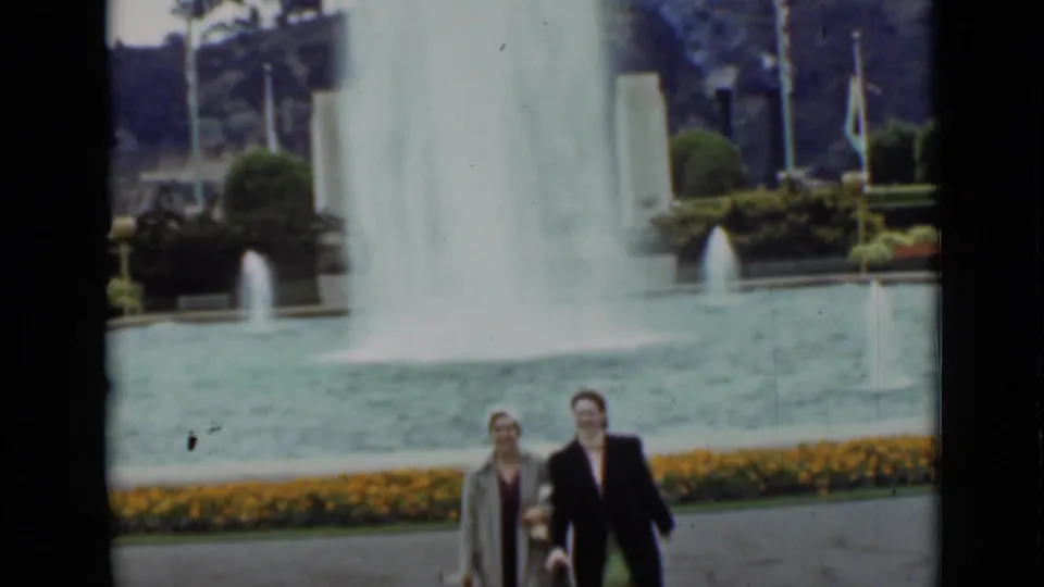 Two women in coats and hats walk in the lower frame while a tall fountain spray rises behind them in the San Francisco park scene.