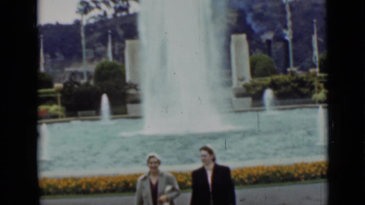 The women pass across the foreground in front of the fountain, with orange flowers, flagpoles, shrubs, and trees visible behind them.