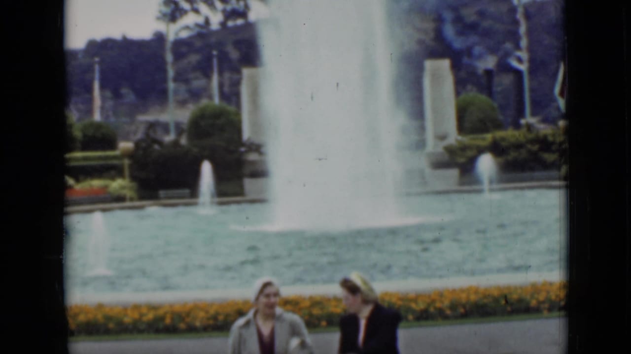 Two women near a fountain pool with flower beds and trees in the background.