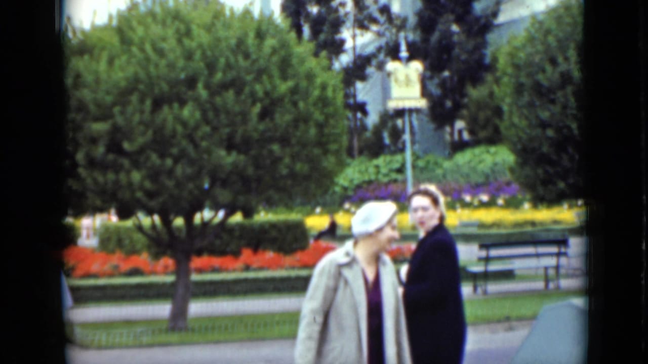 Two women in coats stand near colorful flower beds, a tree, benches, and a park sign.