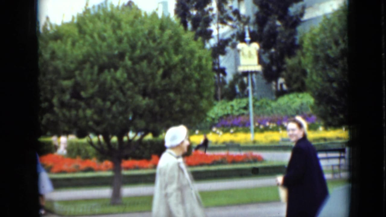 Two women in period outerwear move past manicured hedges and colorful flower beds in 1937 San Francisco home-movie footage.