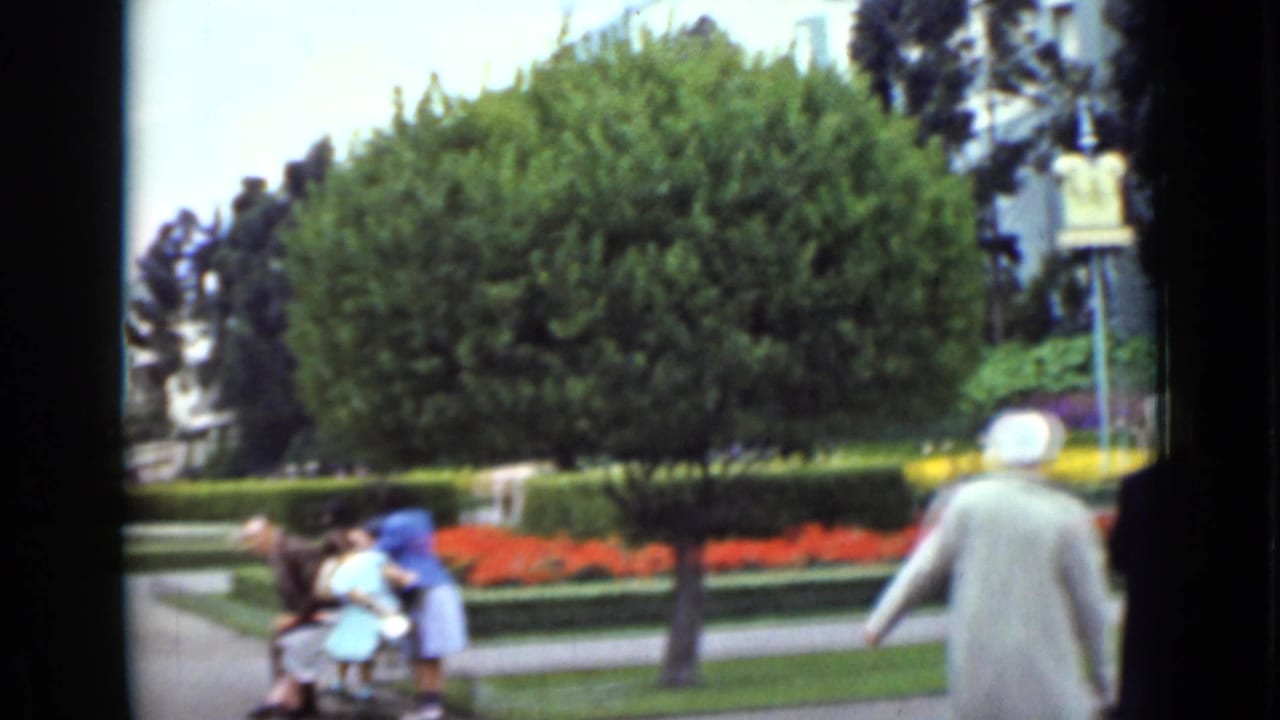 A woman in a light coat walks away through a landscaped San Francisco park as visitors gather near the path and flower beds.