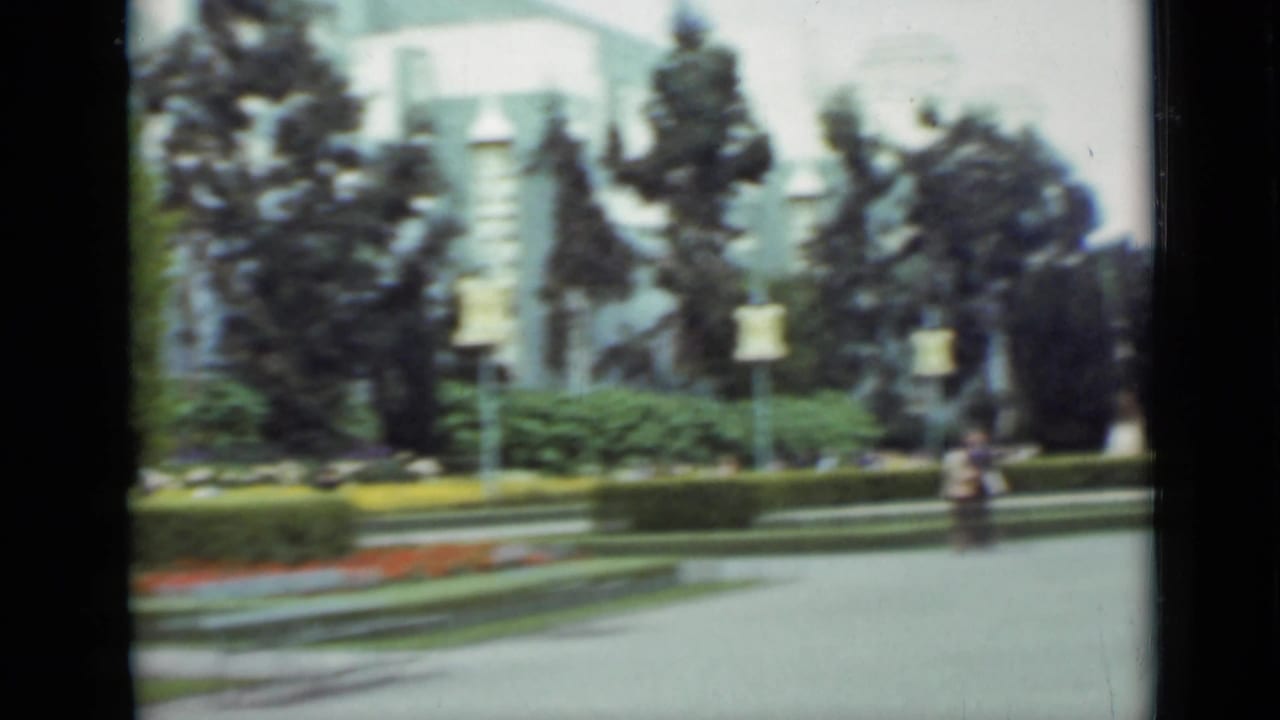 Soft-focus park path with flower beds, trees, and a distant seated figure.