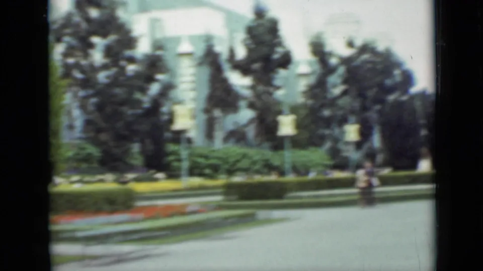 1937 San Francisco home-movie frame showing a landscaped park path, colorful flower beds, trees, and a distant figure near the center.