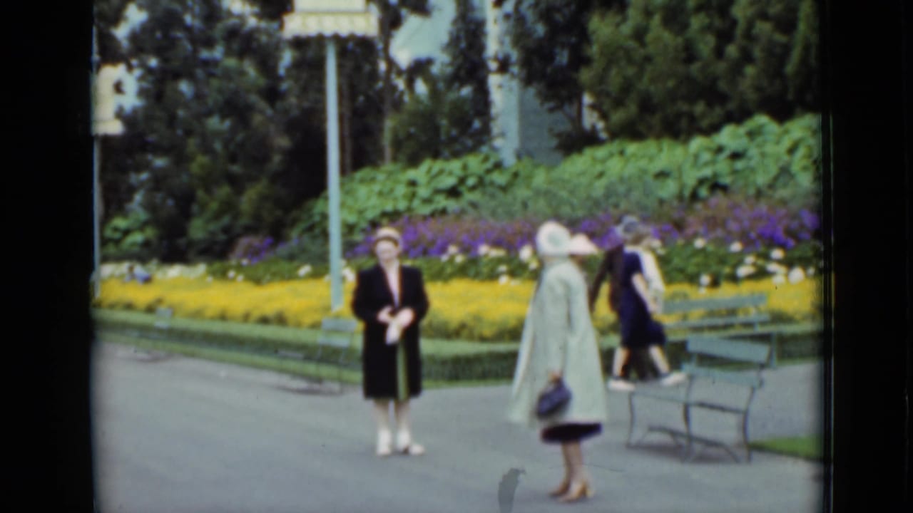 1937 San Francisco frame of two well-dressed women in coats and hats beside benches, lampposts, and yellow and purple flower beds.