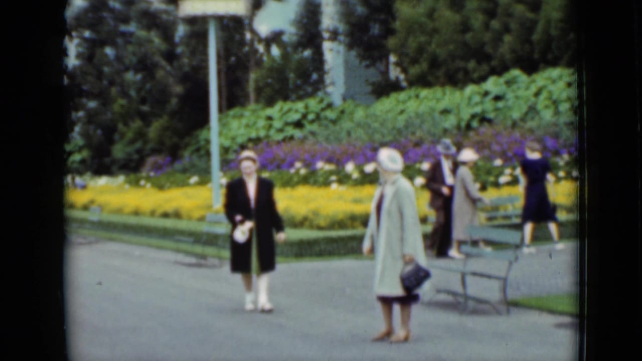 1937 San Francisco home-movie still showing women in formal coats and hats moving through a landscaped park with benches and flower beds.