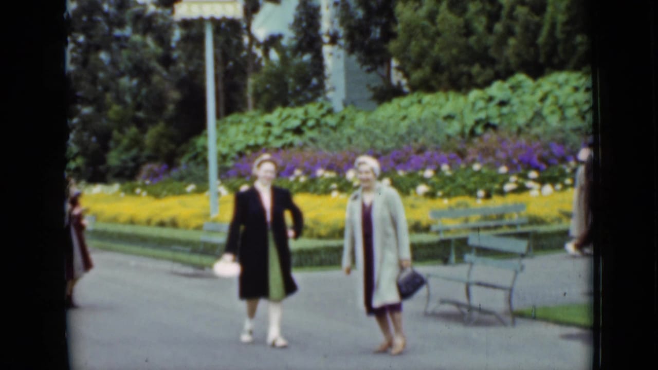 1937 San Francisco frame of two women walking toward the camera in a landscaped park, with benches, trees, and bright flower beds behind them.