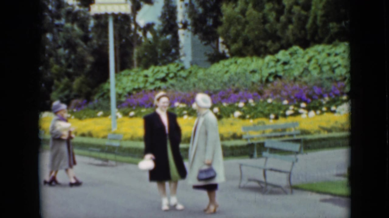Two women in coats and hats stand on a park path near flowerbeds and benches.
