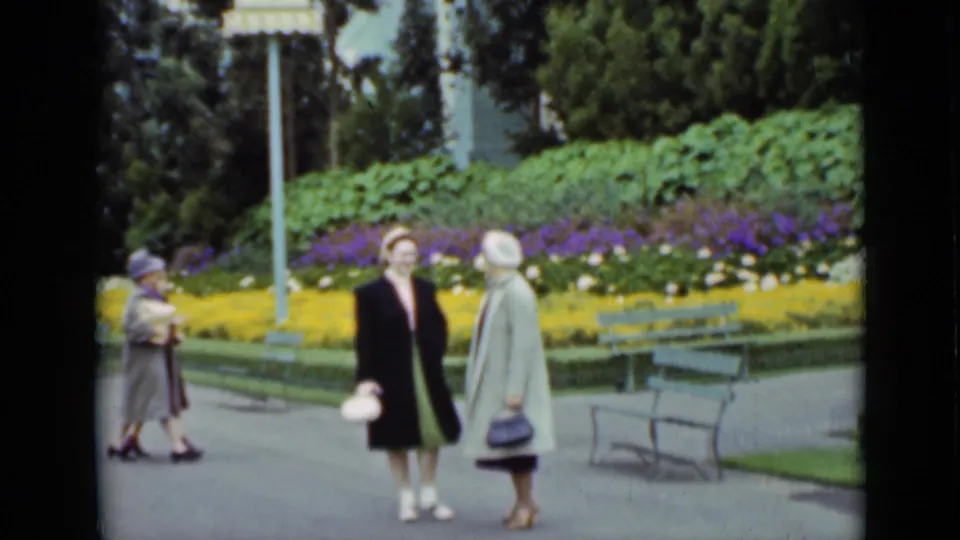 In 1937 San Francisco park footage, two women in formal coats and hats pause on a paved path beside bright flowerbeds and benches.