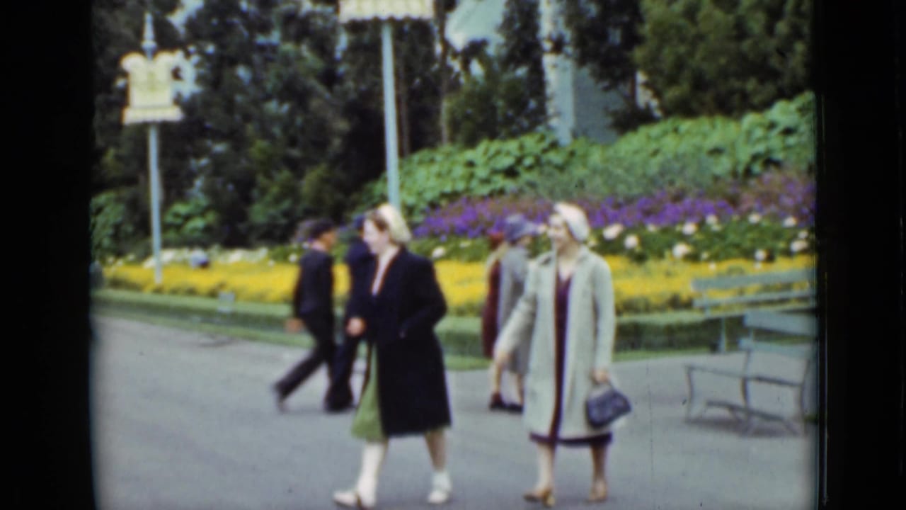 Two women in coats and hats walk through a San Francisco park in 1937, with benches, flowerbeds, lamp posts, and passing visitors nearby.