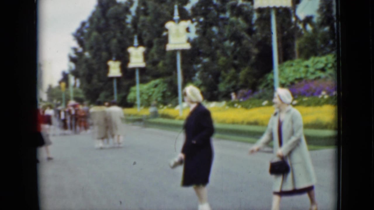 The women continue along a landscaped San Francisco park path, with yellow and purple flowerbeds, lamp posts, and distant pedestrians visible.