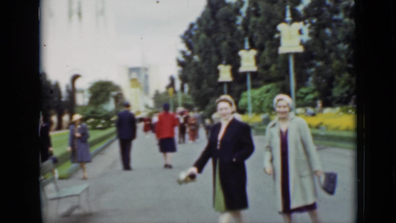 Two women in dress coats and hats approach the camera in 1937 San Francisco park footage, framed by flowerbeds, benches, and distant walkers.