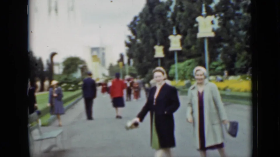 Two women in dress coats and hats approach the camera in 1937 San Francisco park footage, framed by flowerbeds, benches, and distant walkers.