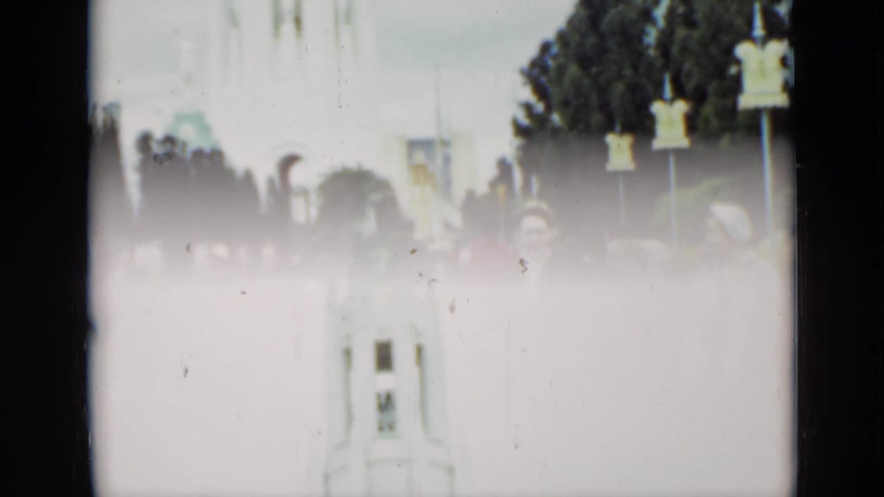 Pedestrians in a square with a pale tower or monument and trees in the background.