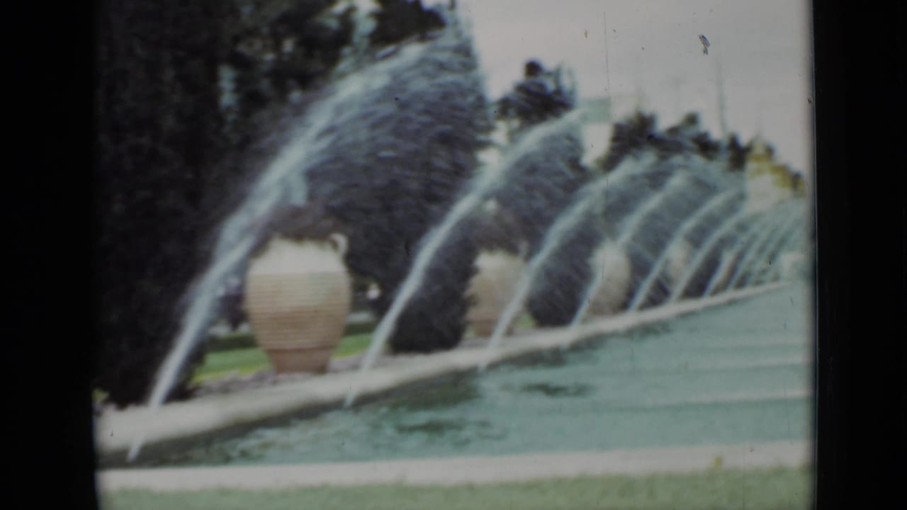 Long fountain with arcing water jets, urn planters, hedges, and trees.