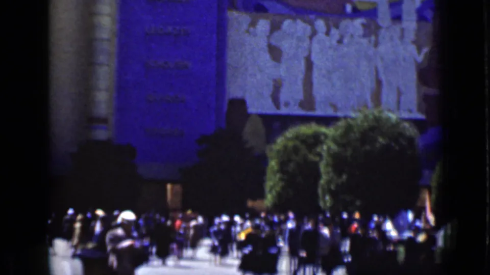 A distant group of San Francisco park visitors stands near a large wall with an indistinct mural or bas-relief and trimmed trees.