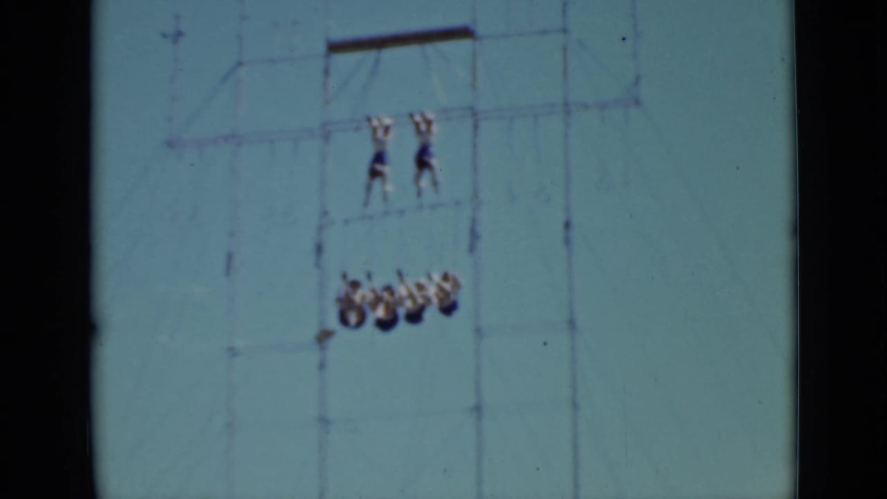 Acrobats hang from a tall metal aerial rig against a blue sky.