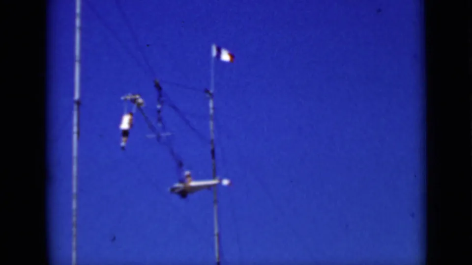 1937 San Francisco frame showing aerial performers on trapeze rigging against a saturated blue sky, with a small tricolor flag on a pole.