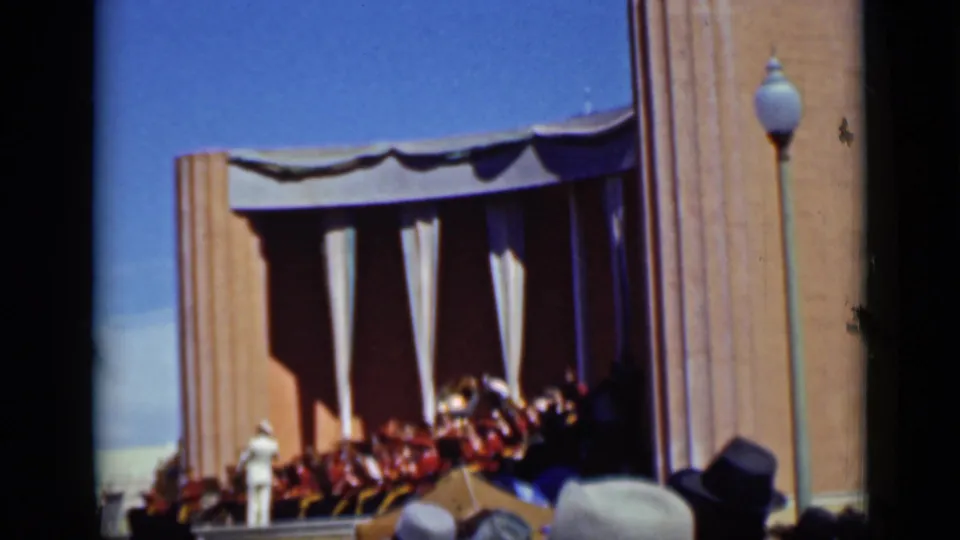 1937 San Francisco home-movie frame showing musicians on an outdoor bandshell stage, with spectators and a lamp post in view.