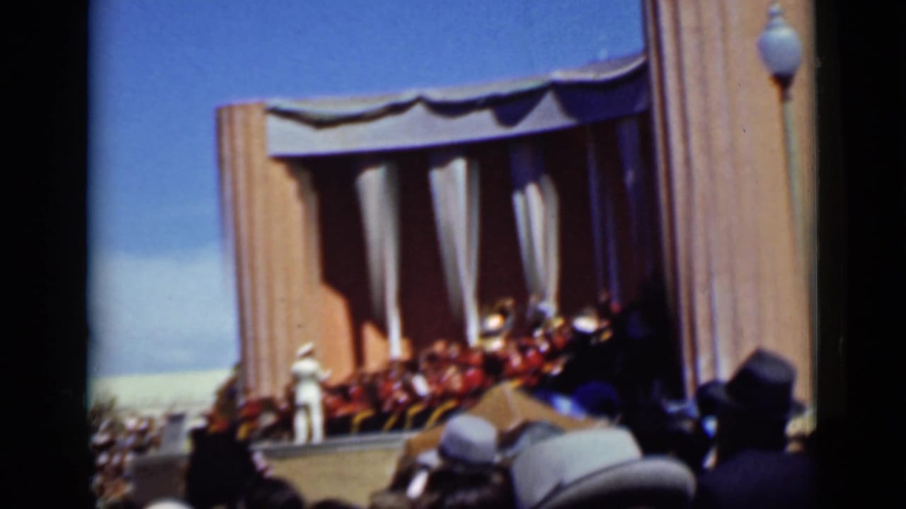 Crowd watches a curtained outdoor stage with a figure in white at the side.