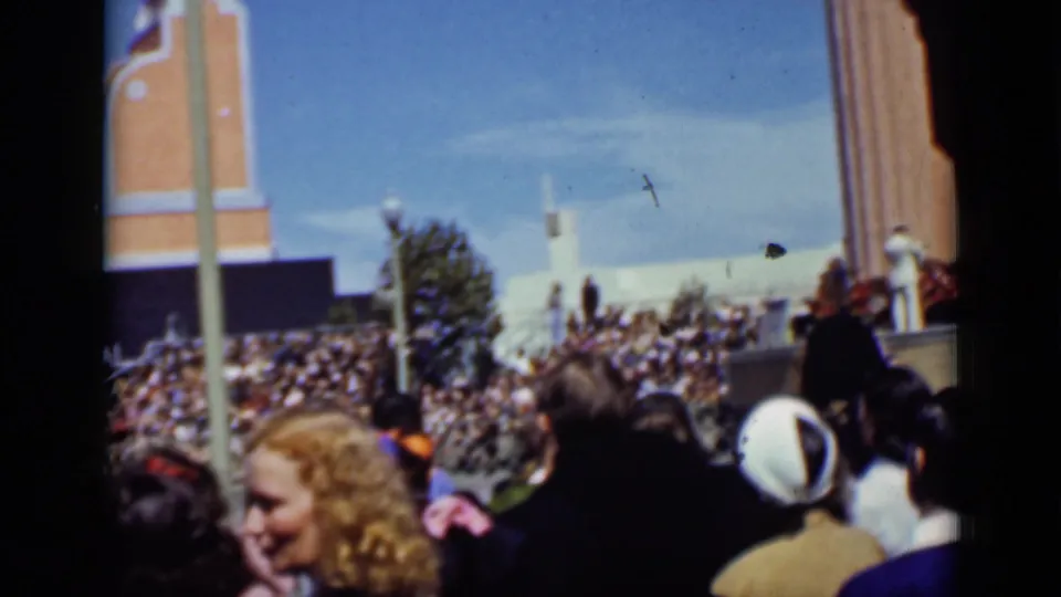 1937 San Francisco frame with dense grandstand seating, foreground audience members, light poles, and a brick tower-like structure under blue sky.
