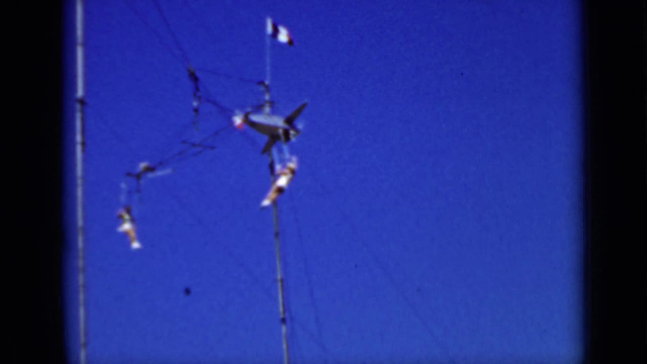 Distant trapeze performers suspended between tall poles against a blue sky.