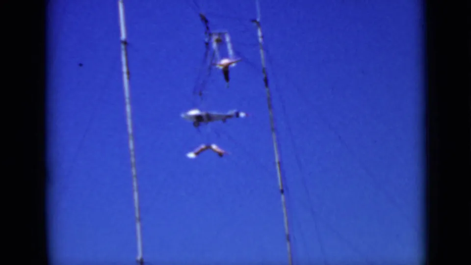 In 1937 San Francisco home-movie footage, two aerial performers hang and swing near tall circus rigging against a vivid blue sky.