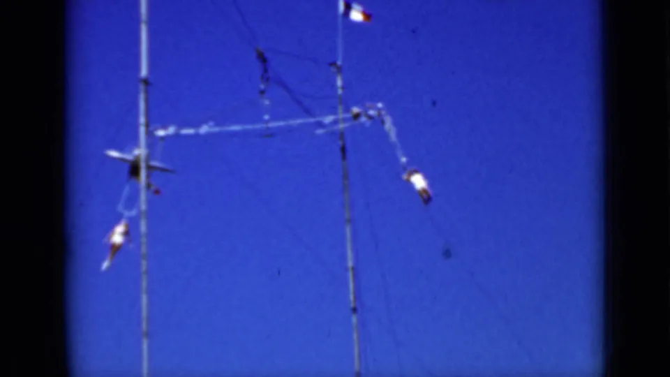 The 8mm frame shows two performers separated on the aerial apparatus, one extending outward while another hangs below the rigging.
