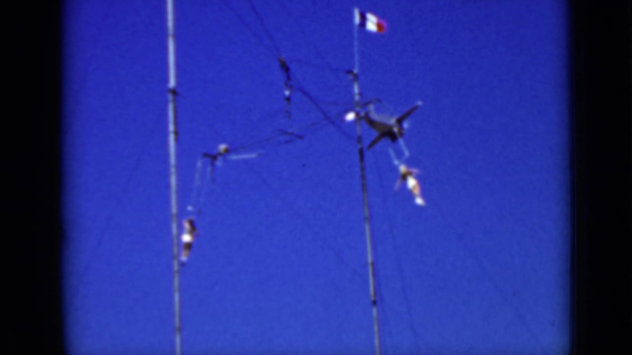 Two aerial performers on a high trapeze rig against a blue sky.