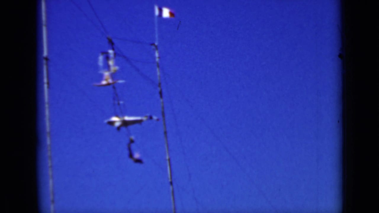 Two trapeze performers swing near tall poles and wires beneath a small tricolor flag.