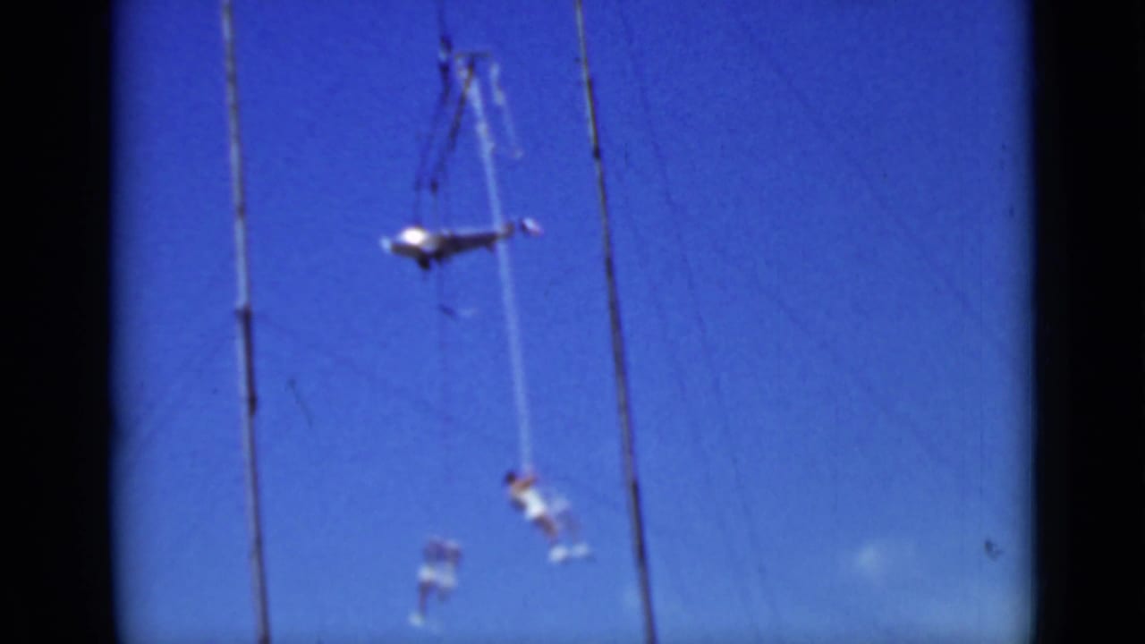 Two aerial performers suspended from ropes near tall rigging poles against a blue sky.