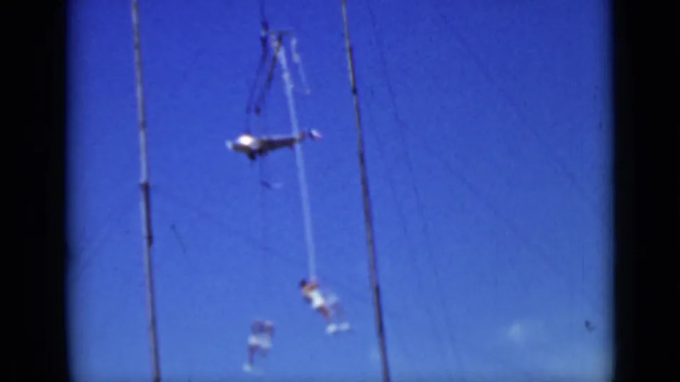 1937 San Francisco 8mm frame showing two distant aerial performers swinging from ropes beside tall rigging poles under a clear blue sky.