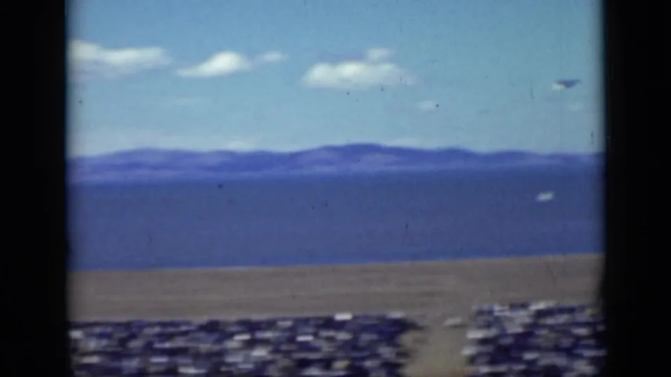 1937 San Francisco 8mm frame showing a distant suspended performer above an open area with hills or water and parked vehicles beyond.