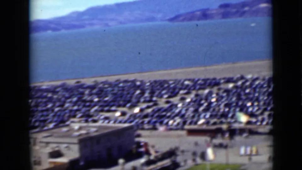 Elevated 1937 San Francisco waterfront frame showing a crowded car lot, small structures, bay water, and distant hills.