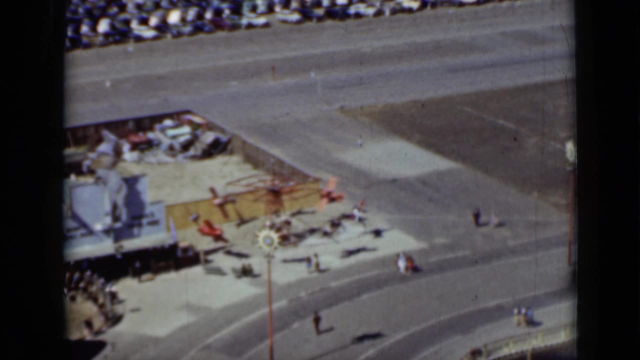 High-angle view of a toy-plane amusement ride near walkways, crowds, and parked cars.