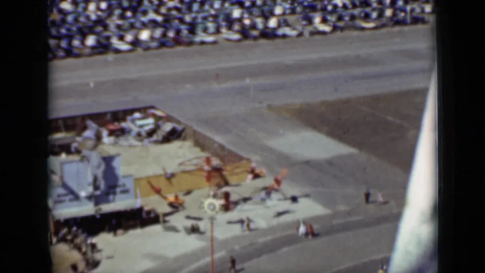 High-angle 1937 San Francisco frame with colorful toy planes on a ride, visitors walking nearby, and parked cars beyond the road.