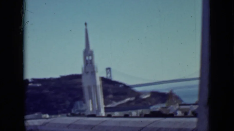 1937 San Francisco home-movie view of a tall white tower with bridge towers, bay water, hillside, and rooflines behind it.