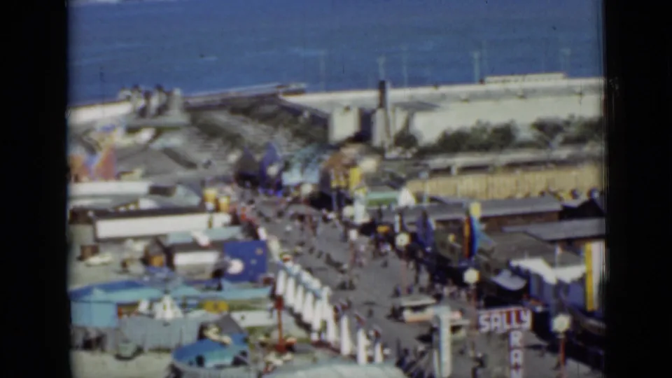 Soft-focused 1937 San Francisco boardwalk frame showing a busy amusement promenade, ocean background, and visible entertainment signage.