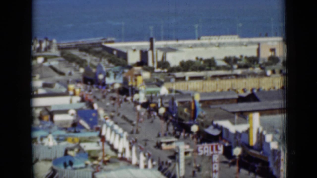 Elevated view of a boardwalk amusement area near the ocean with crowds, ride structures, and partial signage.